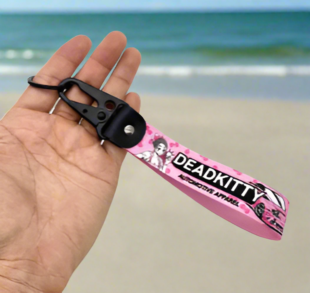 Pink DEADKITTY lanyard with cartoon character, held against beach backdrop.