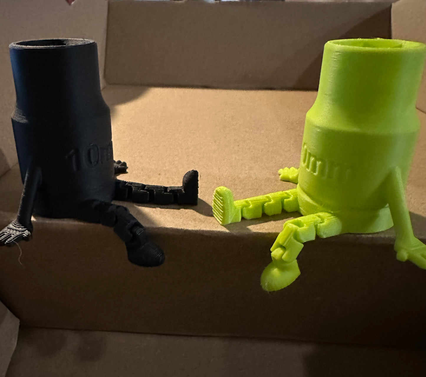 Black and green 3D-printed objects with legs on cardboard surface.