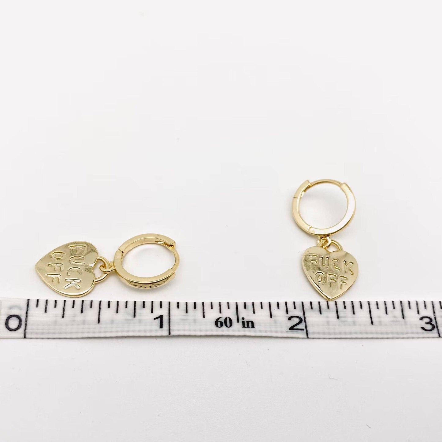 Gold hoop earrings with FUCK OFF heart charms next to a ruler.