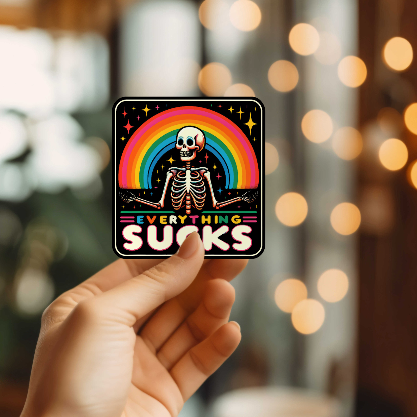 Hand holding a sticker with a skeleton and rainbow, Everything Sucks text.