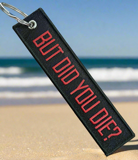Black keychain with red text BUT DID YOU DIE on sandy beach background.