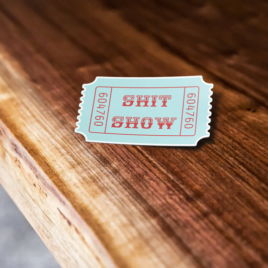 Light blue ticket-shaped sticker with SHIT SHOW text on wooden surface.