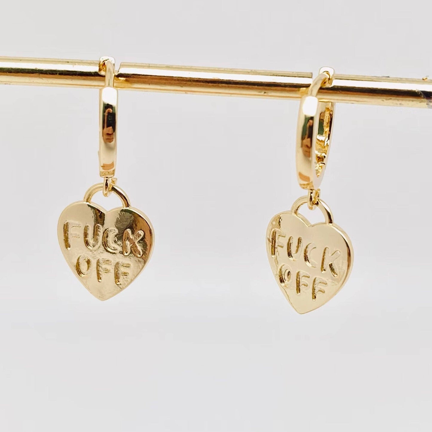 Gold heart-shaped earrings with FUCK OFF engraved on them.