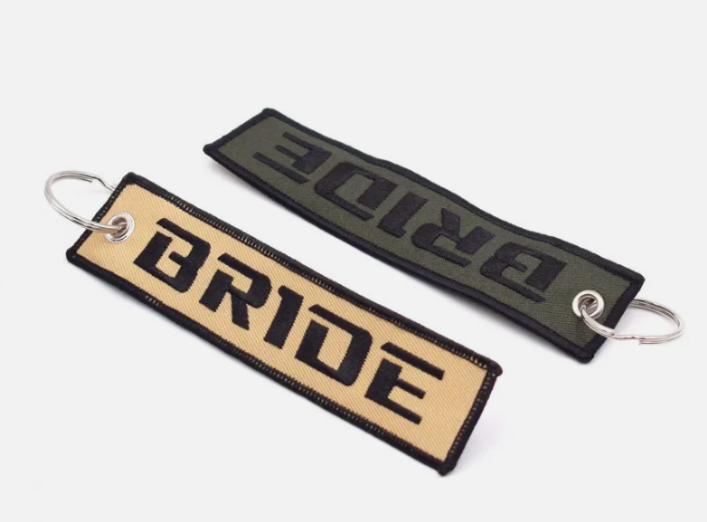 Two BRIDE embroidered keychains in beige and olive green.