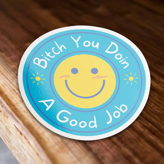 Circular sticker with smiley face and text Bitch You Doin A Good Job on a wooden surface.