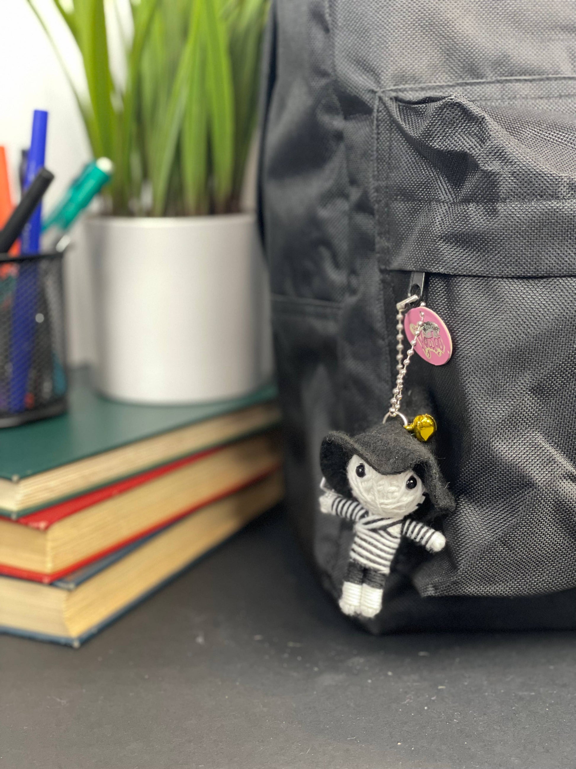 Black backpack with doll charm and pink badge, books and plant in background.
