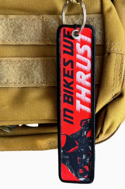 Red and black keychain with IN BIKES WE TRUST text on a tactical bag background.