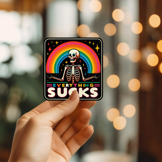 Hand holding a sticker with a skeleton and rainbow, Everything Sucks text.