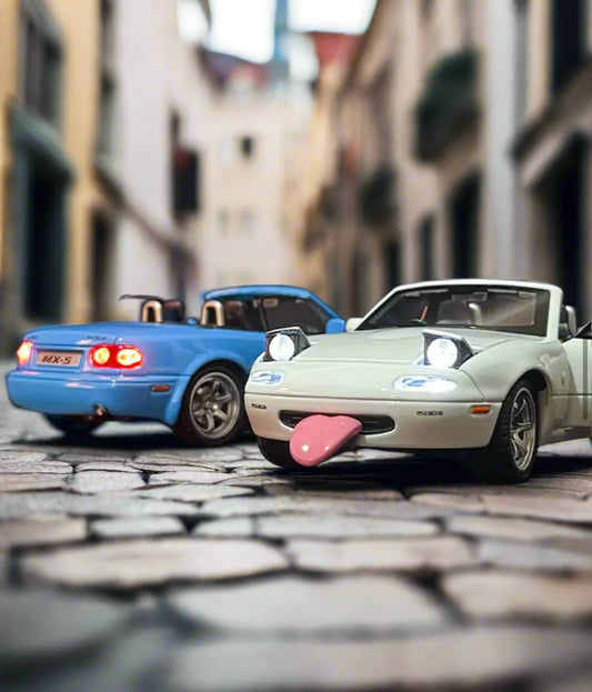 Two toy Mazda MX-5 cars on cobblestone street, one with a pink tongue sticking out.