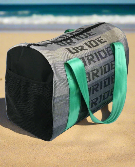 Gray beach bag with green handles and BRIDE text on sand near ocean.