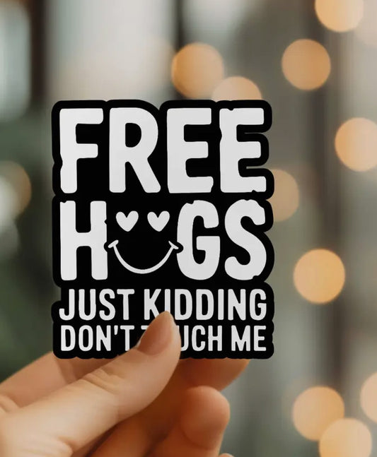 Hand holding a sticker that says FREE HUGS JUST KIDDING DONT TOUCH ME with bokeh lights in the background.