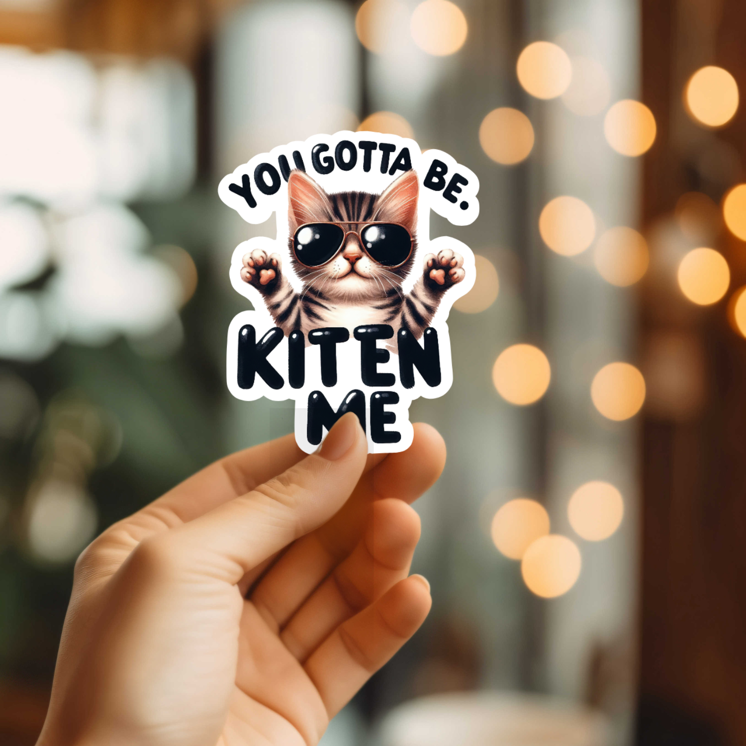 Hand holding a sticker with a cool kitten wearing sunglasses and the text YOU GOTTA BE. KITEN ME.
